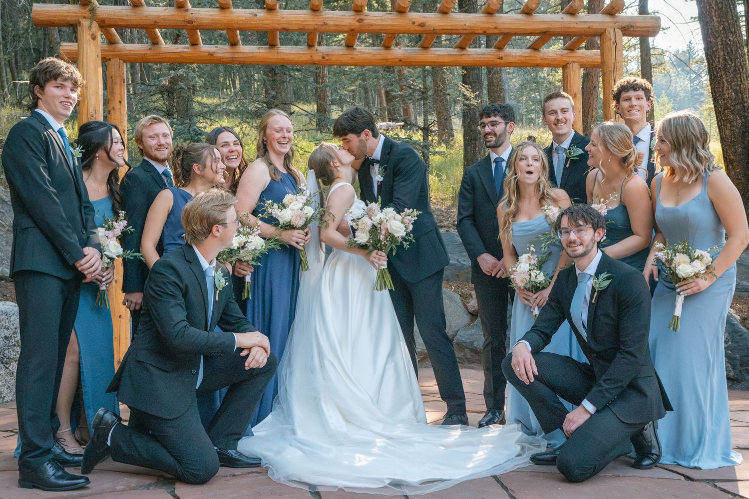 Wedding party poses with kissing bride and groom, bridesmaids in blue dresses and groomsmen in dark suits outdoors.