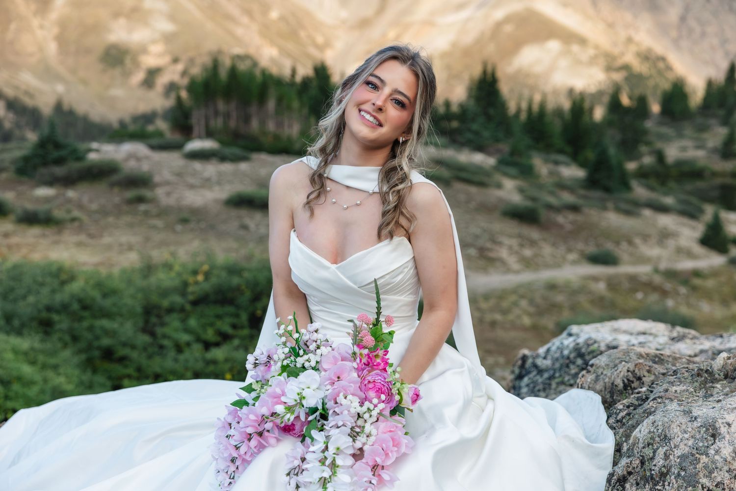 Smiling bride in white strapless gown holding pink floral bouquet, seated on rocks with mountain scenery backdrop.