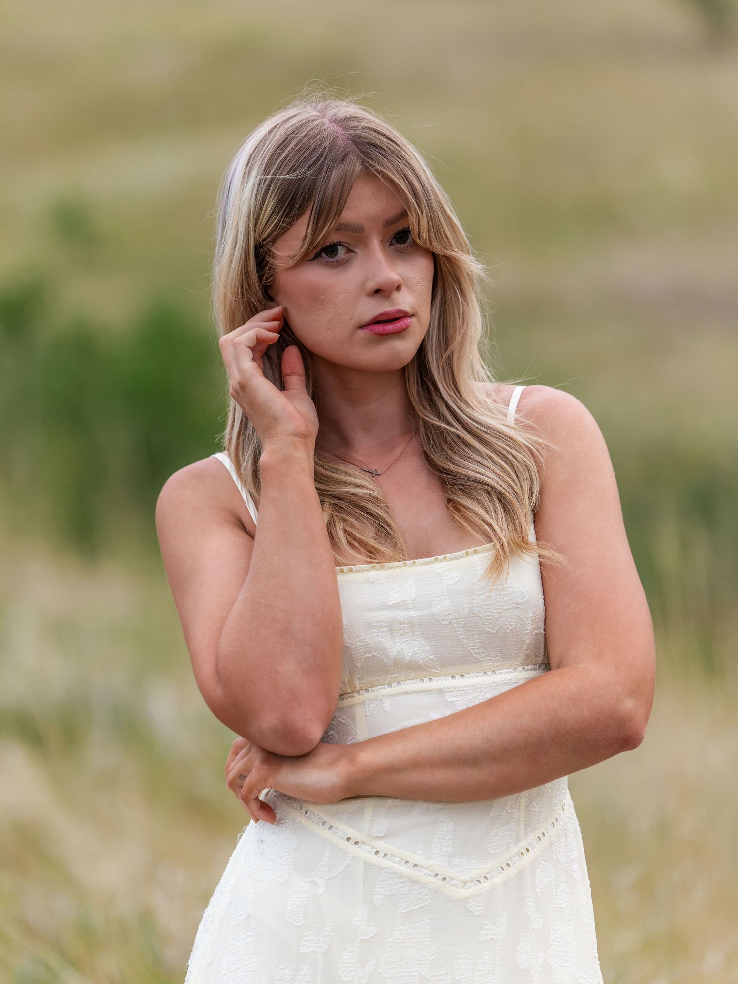 Young woman in white sundress poses thoughtfully in a golden meadow, touching her ear with wavy blonde hair.
