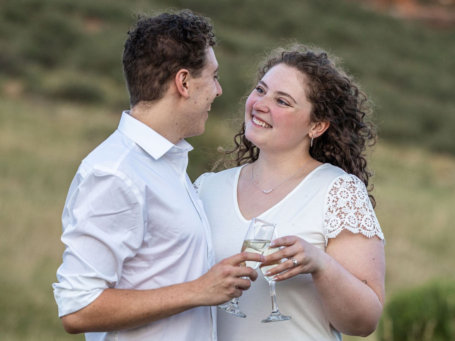 Couple smiling at each other while holding champagne glasses outdoors in a romantic engagement photo.