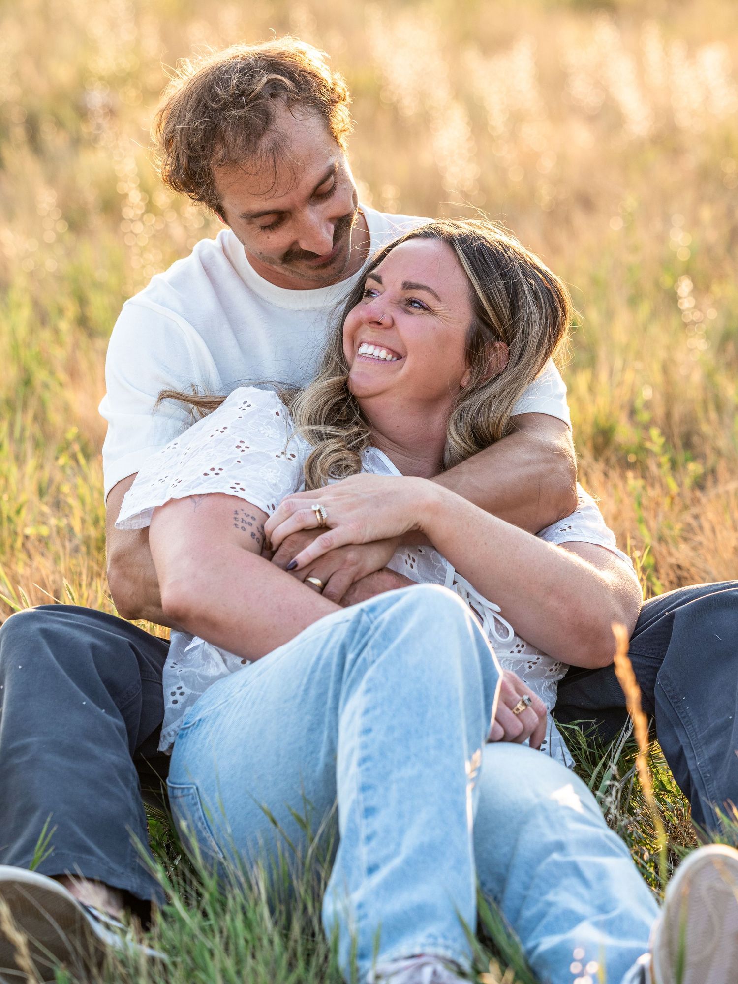 Couple sitting in a golden grass field, laughing and embracing during an outdoor engagement photo session.