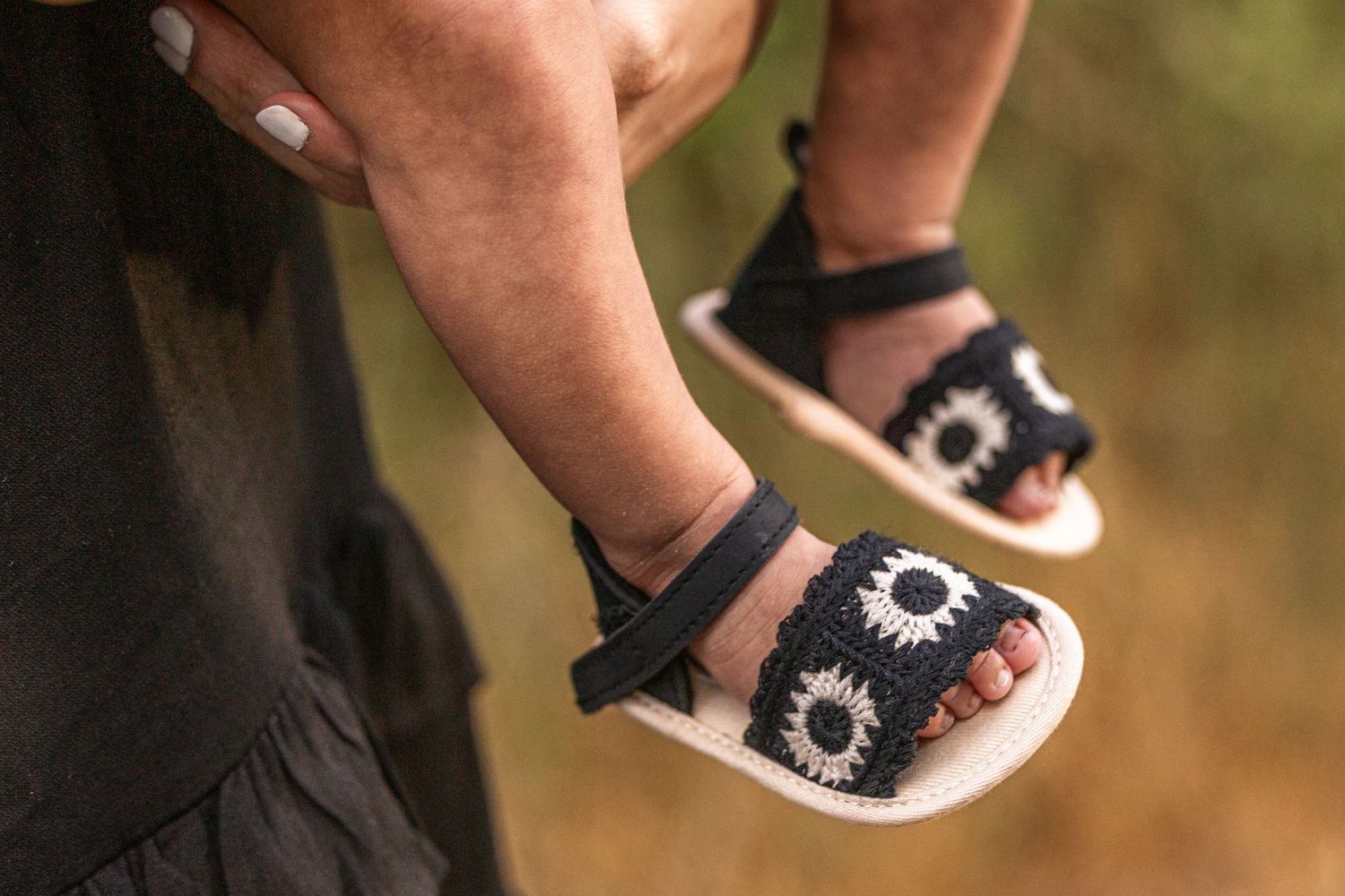 Close-up of a baby wearing black crochet sunflower sandals being held by an adult outdoors.