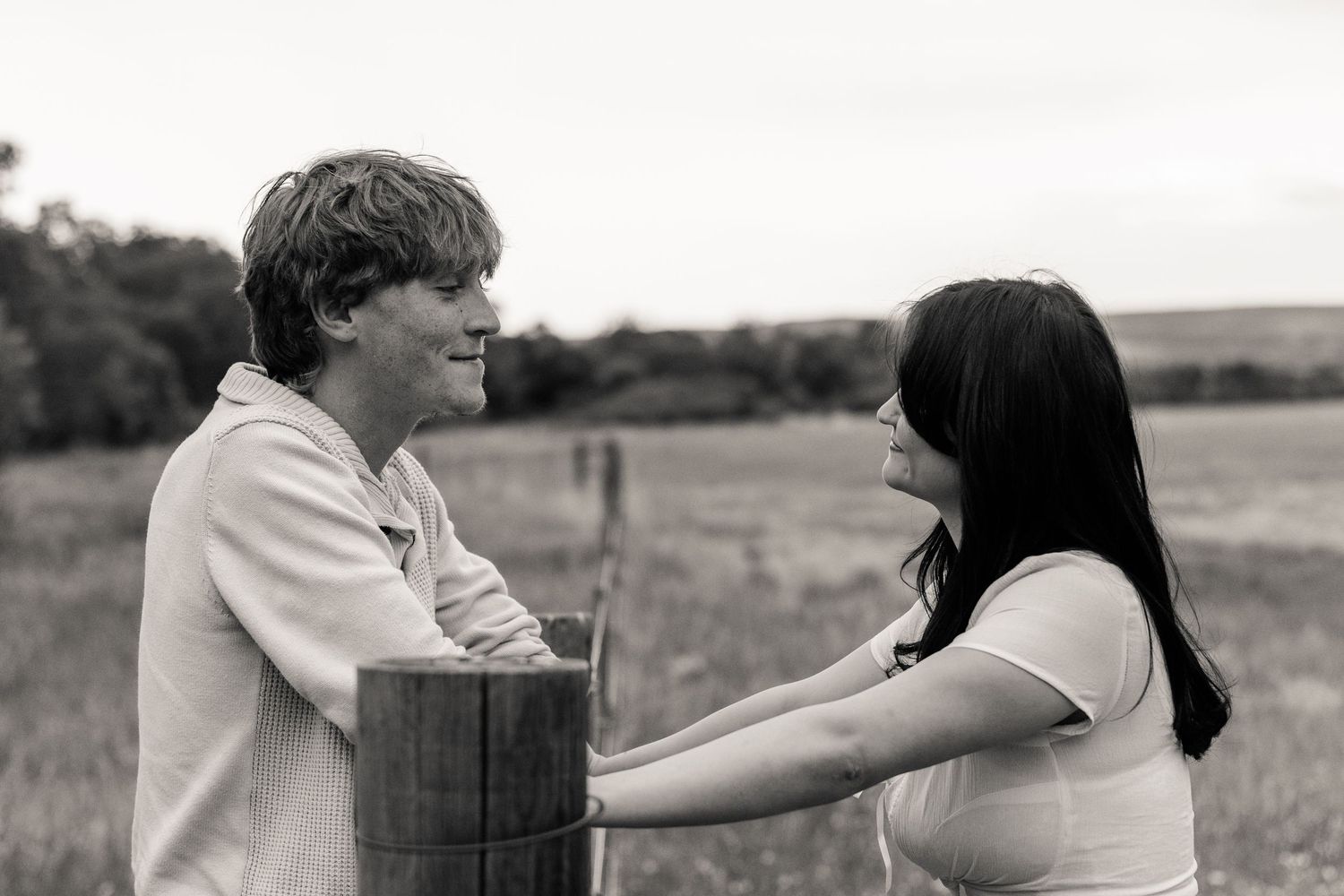 Black and white photo of a young couple sharing a tender moment by a wooden post in a rural field.