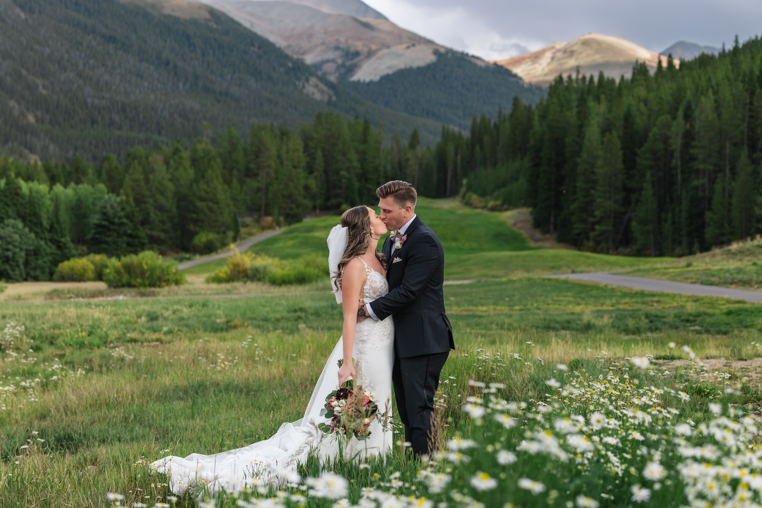 Bride and groom share a romantic kiss in a wildflower meadow with mountain forest backdrop.