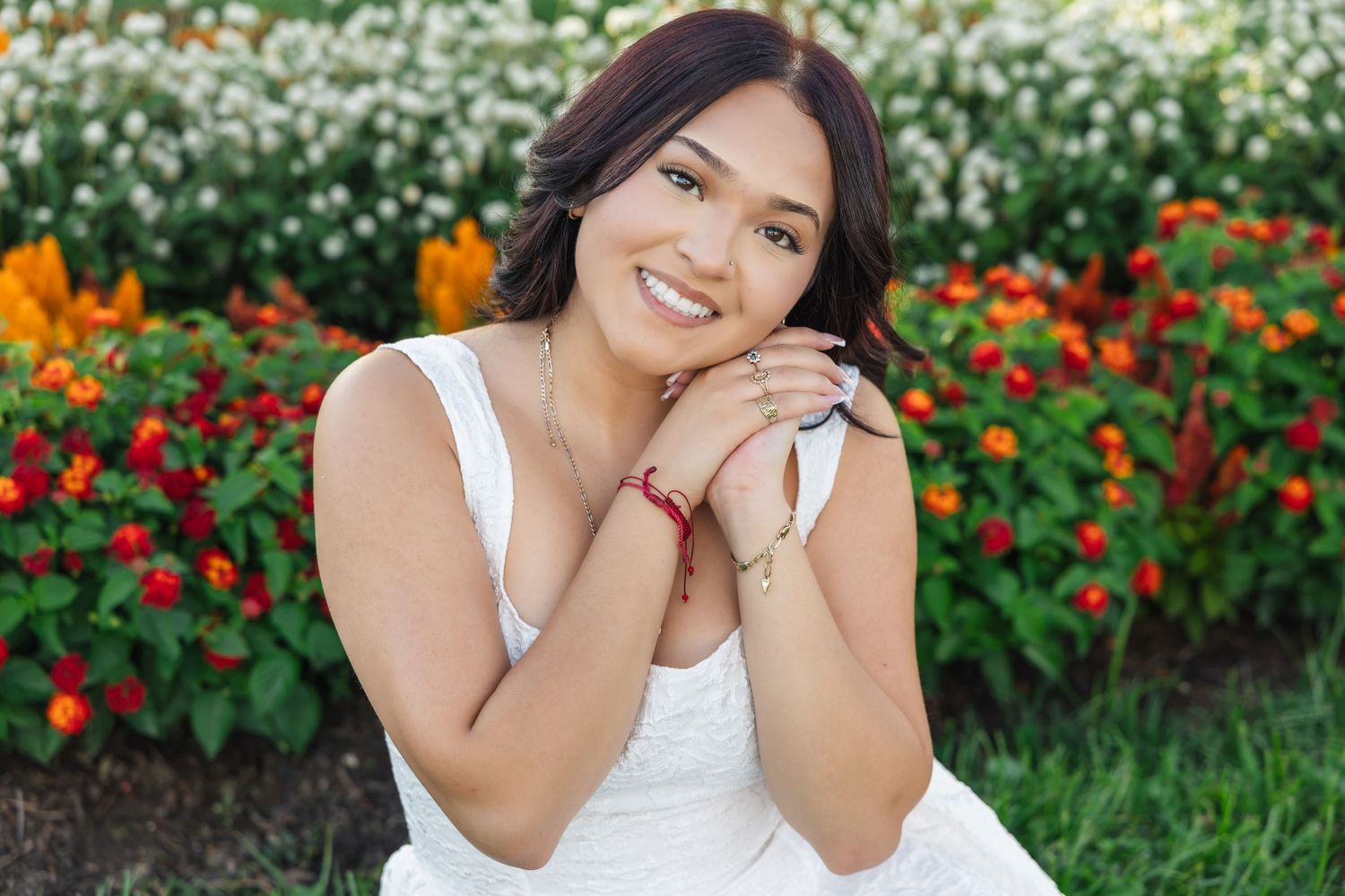 Young woman in white dress smiling in front of colorful flower garden, hands clasped near face.