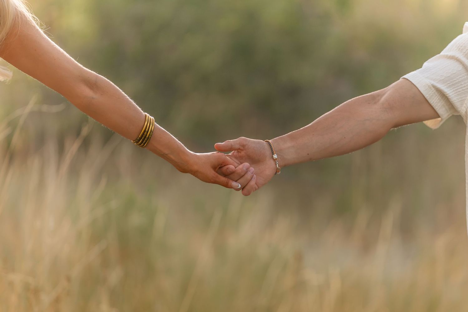 Couple holding hands in a sunlit field, woman wearing gold bracelet, man in white shirt, romantic outdoor setting.