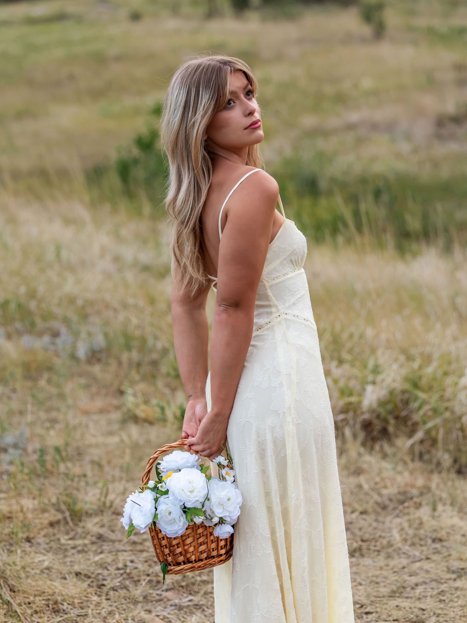 Woman in cream maxi dress holding white flower basket stands in golden field, glancing over shoulder.