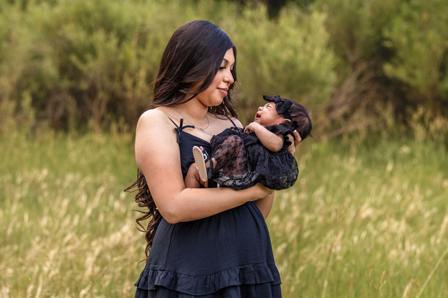A woman in a black dress holds her newborn baby outdoors in a lush green field during a maternity photo shoot.