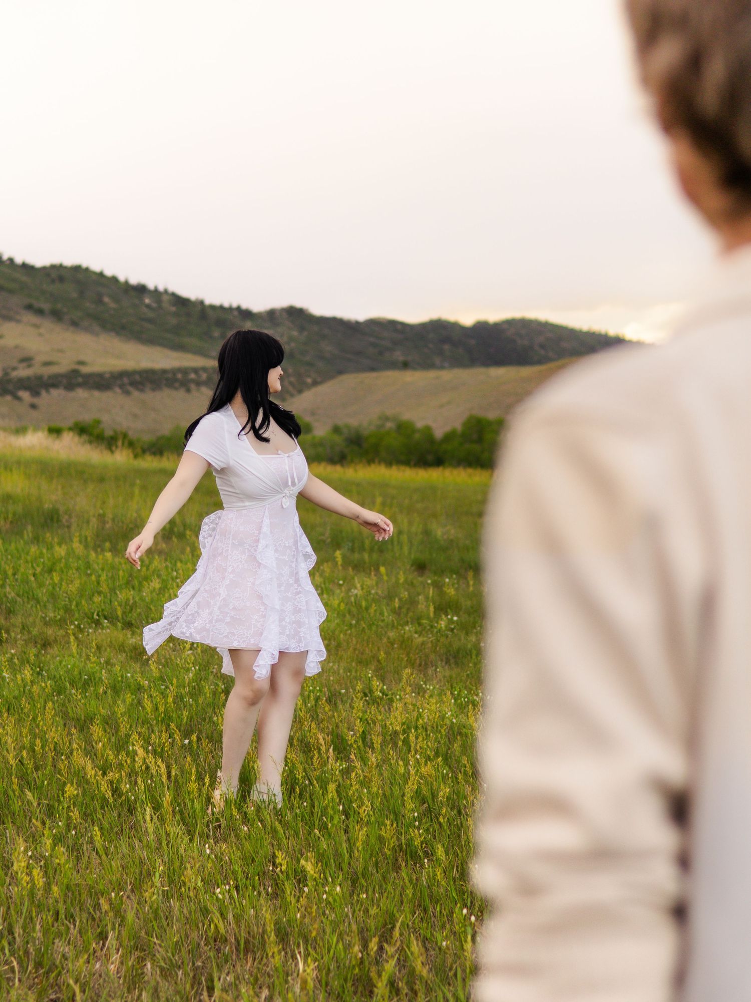 Woman in white dress twirling in green meadow at dusk with rolling hills in background in Littleton, Colorado.