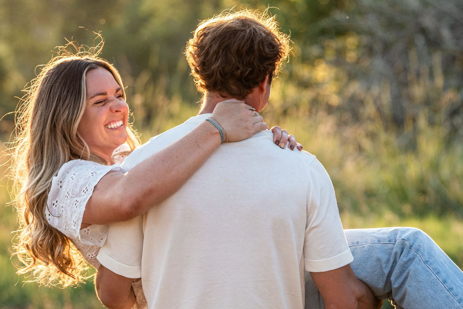 A smiling woman with long hair embraces a man outdoors in golden sunlight, her arm around his shoulder.