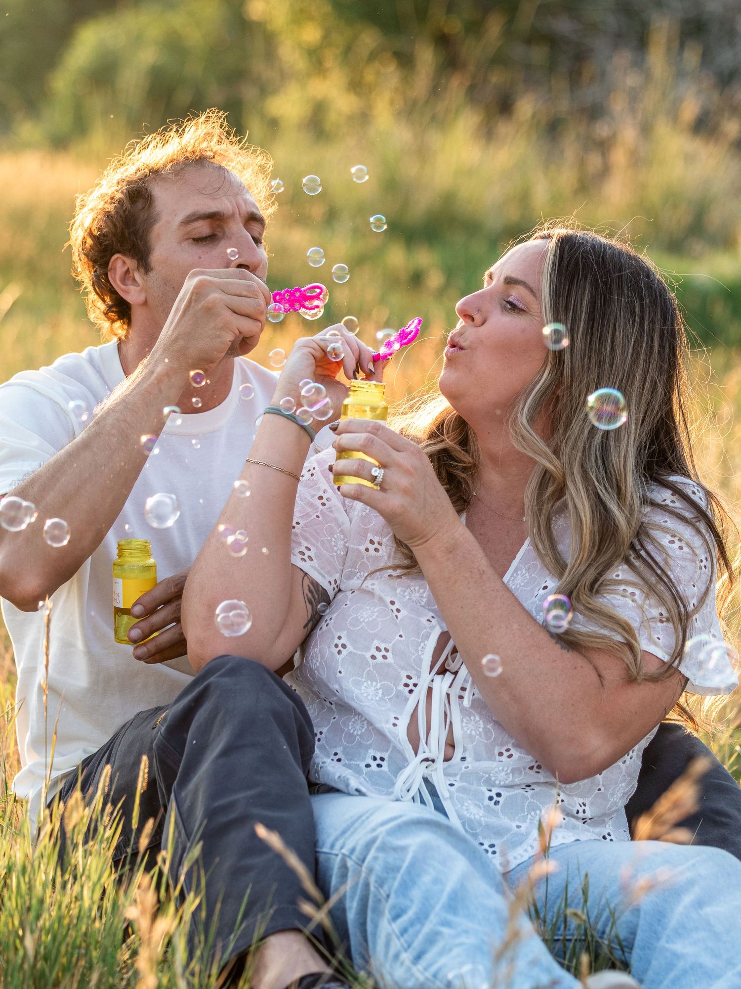Couple sitting in golden grass blowing soap bubbles together in a sunny outdoor field, wearing white outfits.