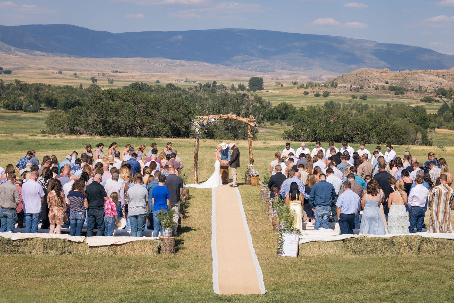 Outdoor wedding ceremony in a scenic mountain valley with guests seated on white chairs along a rustic burlap aisle runner.