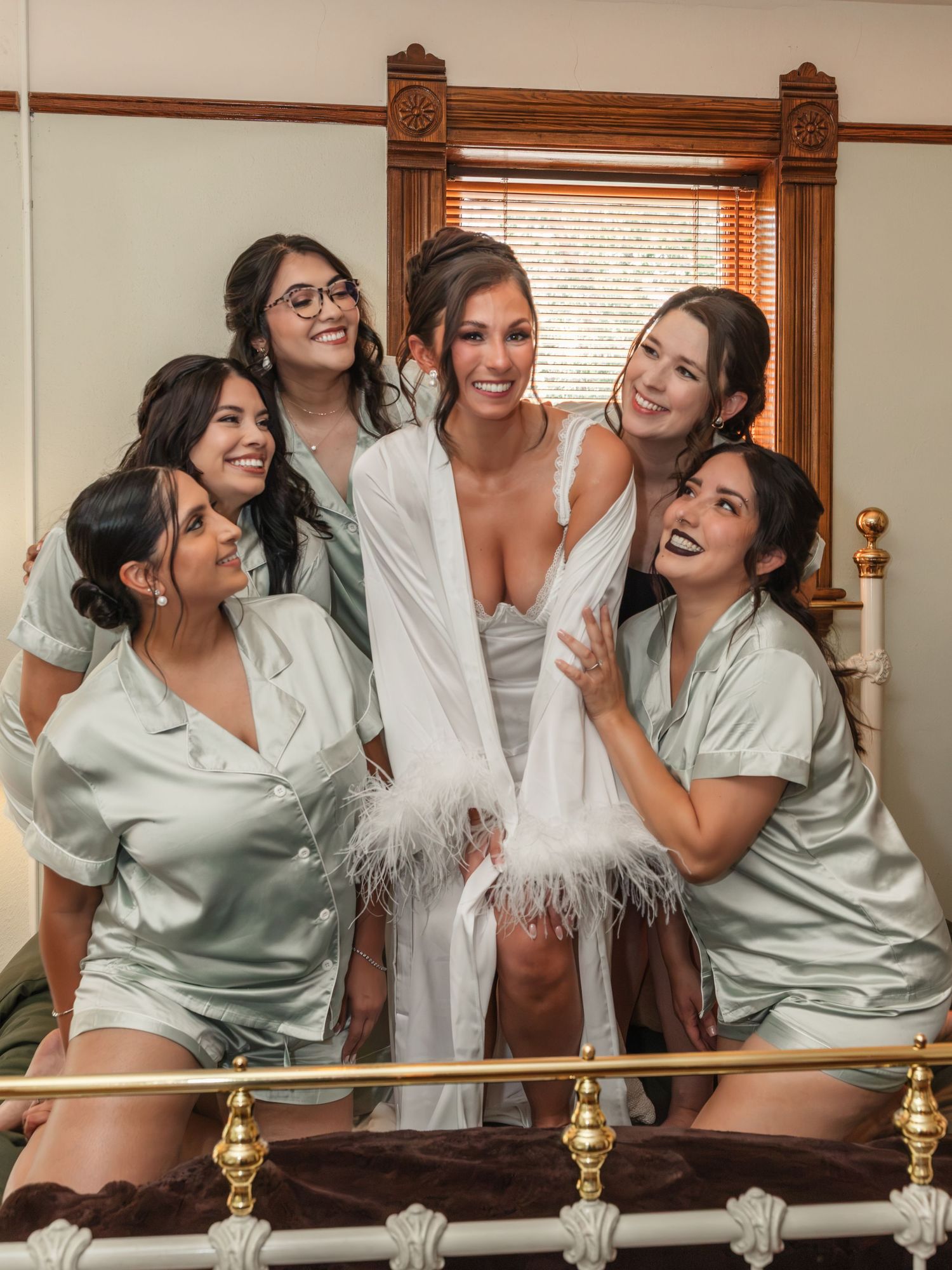 Bride in white feather robe surrounded by bridesmaids in sage satin pajamas laughing together on a bed.