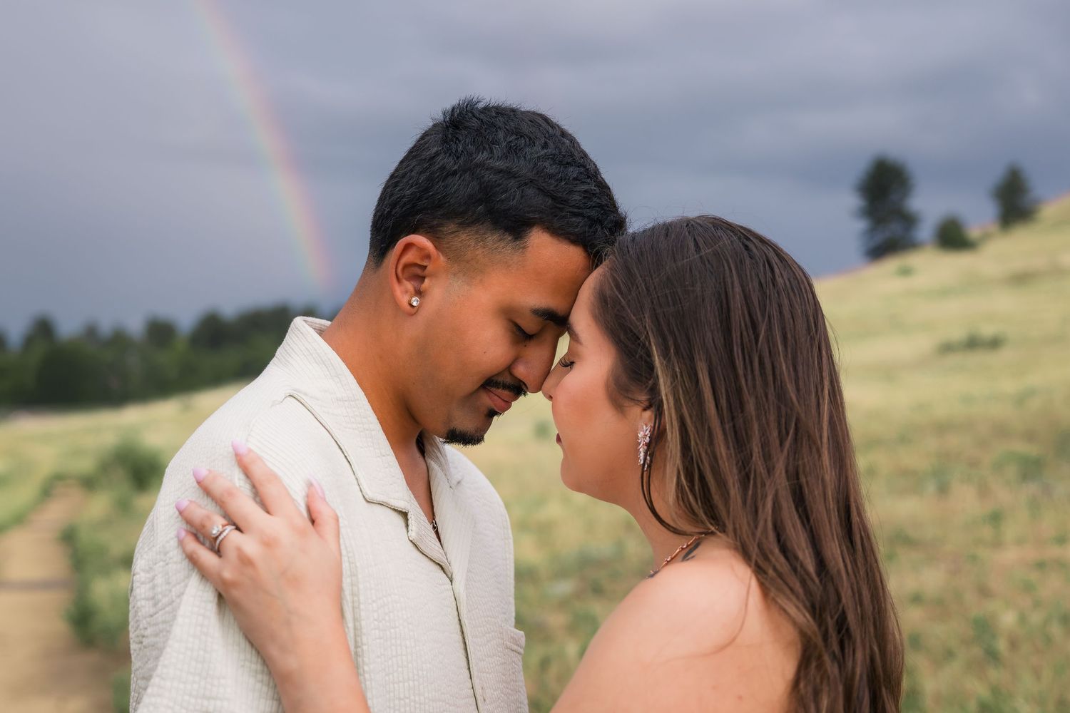 Couple shares a romantic forehead touch outdoors with a vibrant rainbow arching over green hills in the background.