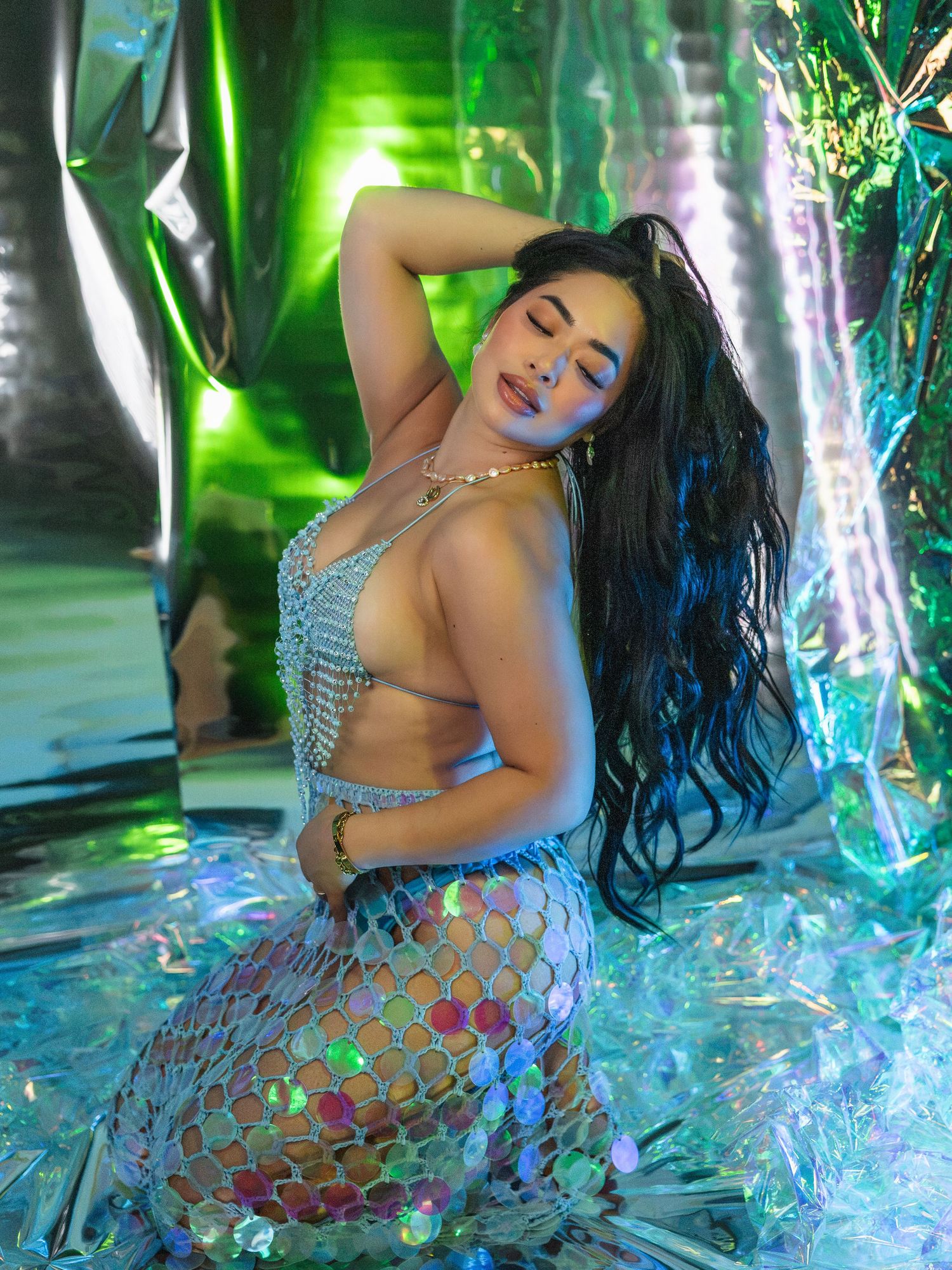 Woman in iridescent mermaid costume posing against holographic green and silver backdrop.