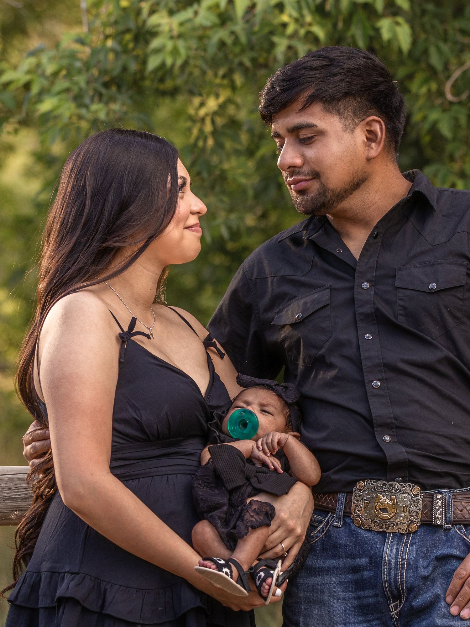 Young Hispanic couple in black outfits gazing at each other while holding a newborn baby outdoors.