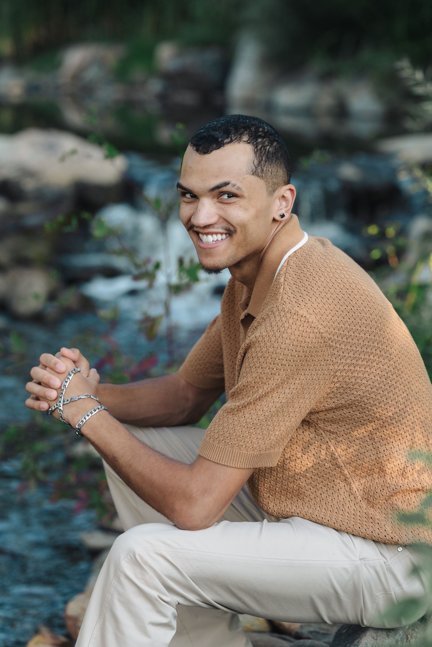Smiling young man in brown knit shirt and khakis sits outdoors near a waterfall, wearing silver jewelry.