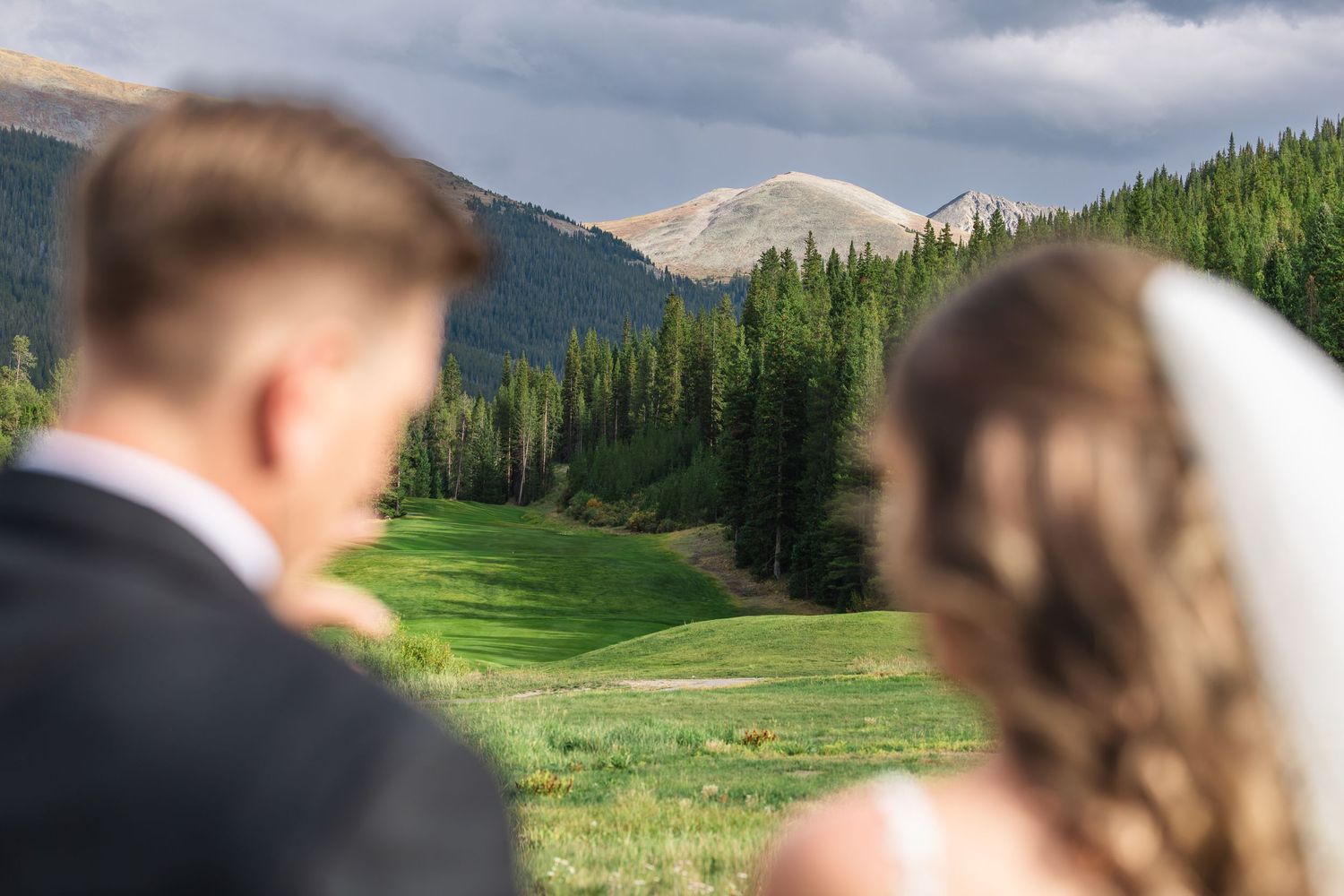 Bride and groom stand together overlooking a lush green mountain valley with pine trees and rocky peaks on their wedding day.