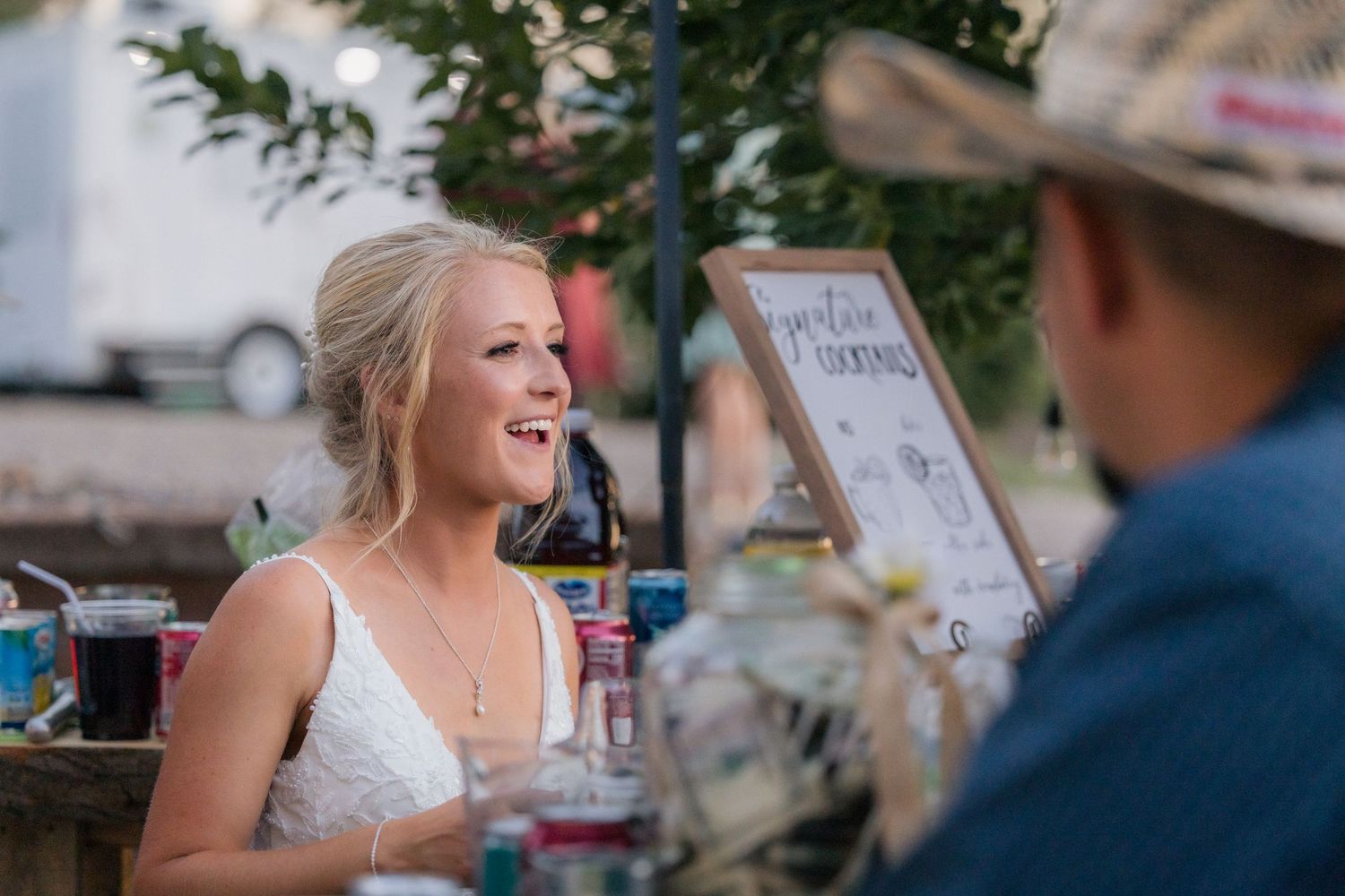 Smiling bride in white dress sits at outdoor wedding reception table, laughing with guest wearing cowboy hat.