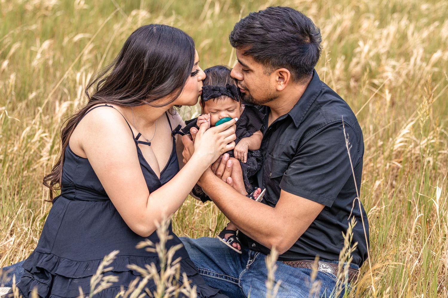 A young family sits in a golden field, parents lovingly kissing their newborn baby outdoors.