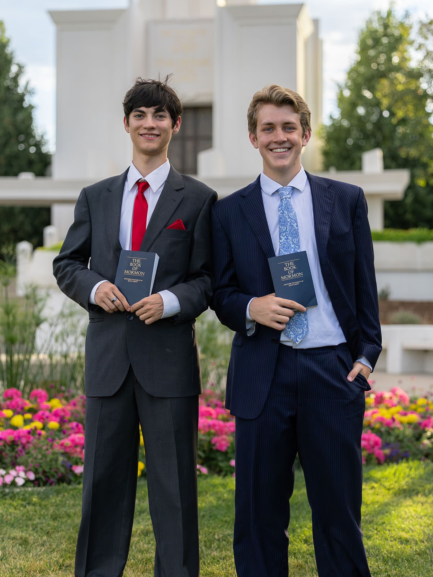 Two young men in suits holding Books of Mormon pose outdoors near a temple with colorful flowers.