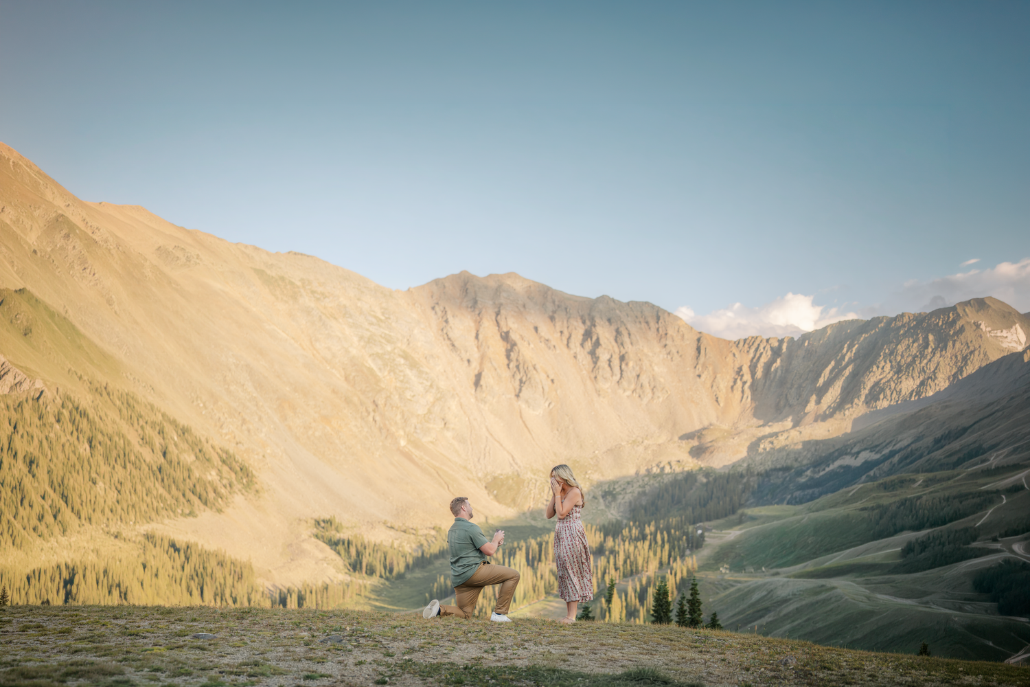 A couple shares a romantic marriage proposal moment on a mountain ridge with stunning valley and peaks backdrop.
