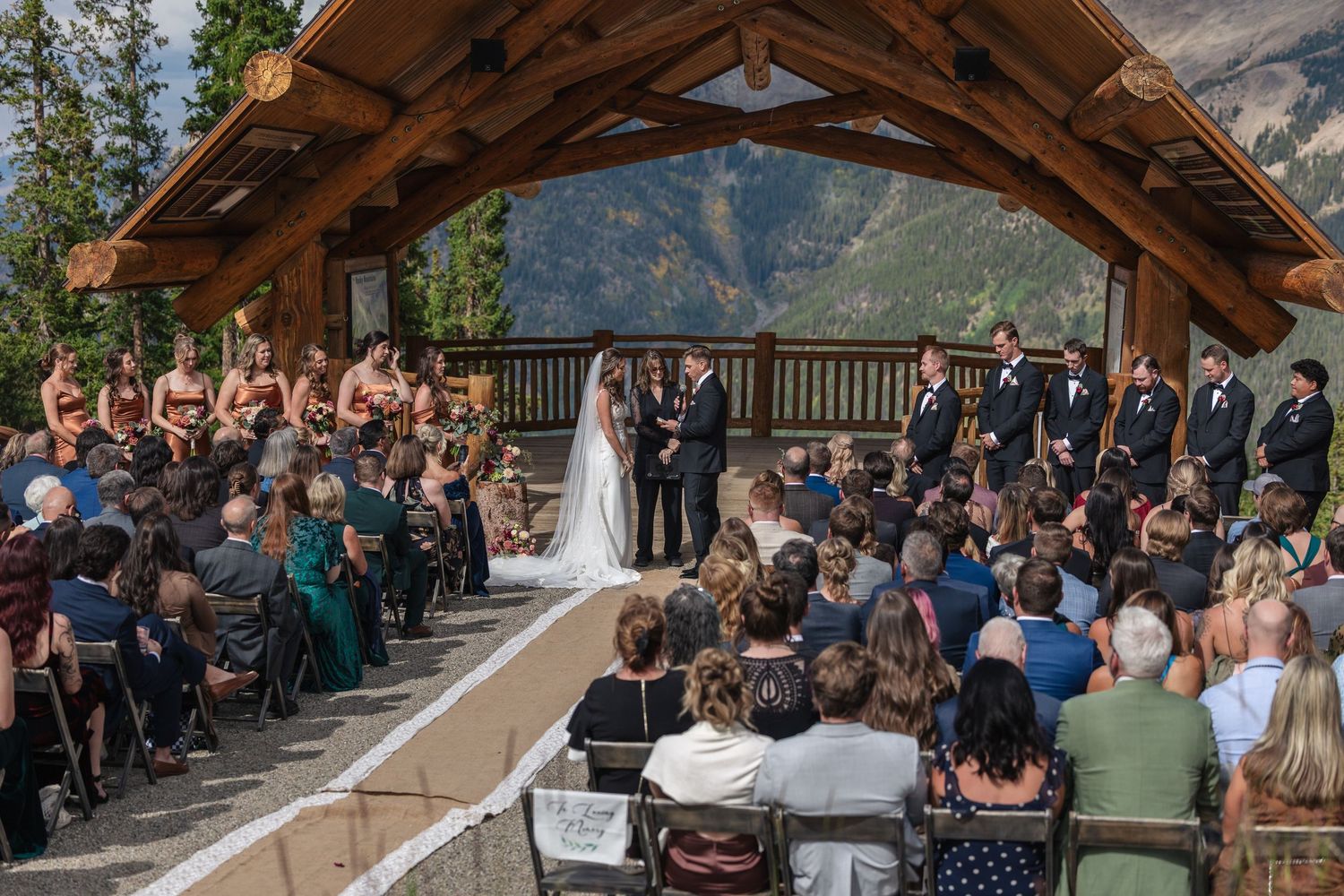Outdoor wedding ceremony under a rustic wooden pavilion with mountain views, featuring bride, groom, and guests.