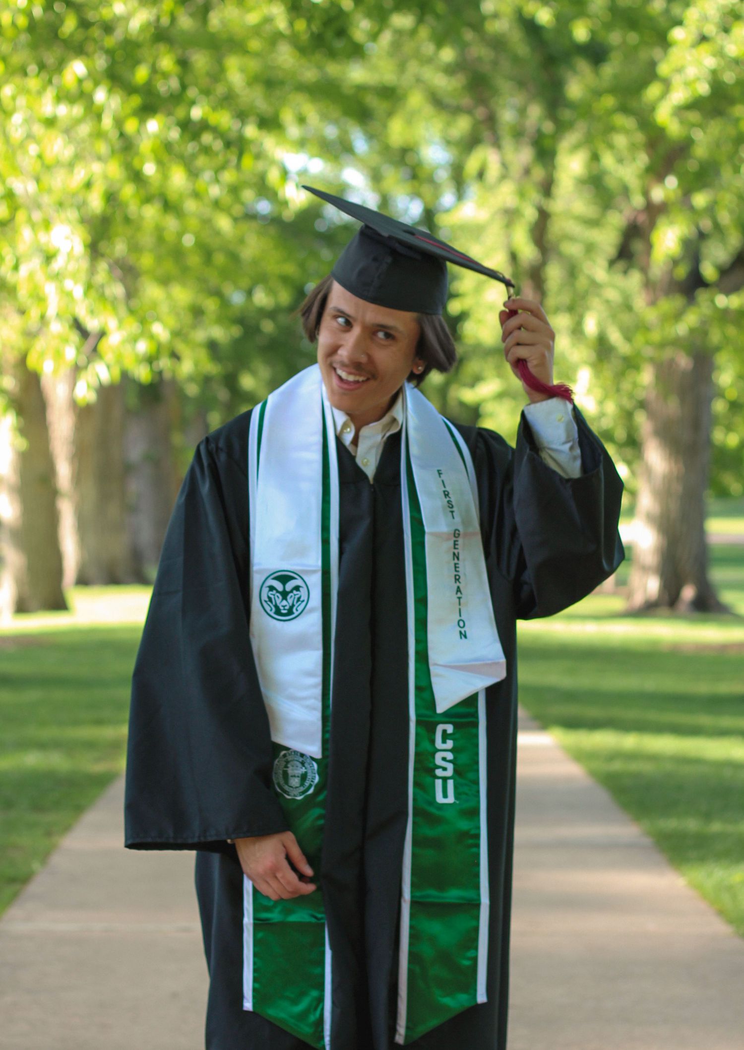 Graduate in black cap and gown with green CSU stole smiles while holding tassel outdoors on tree-lined campus path.
