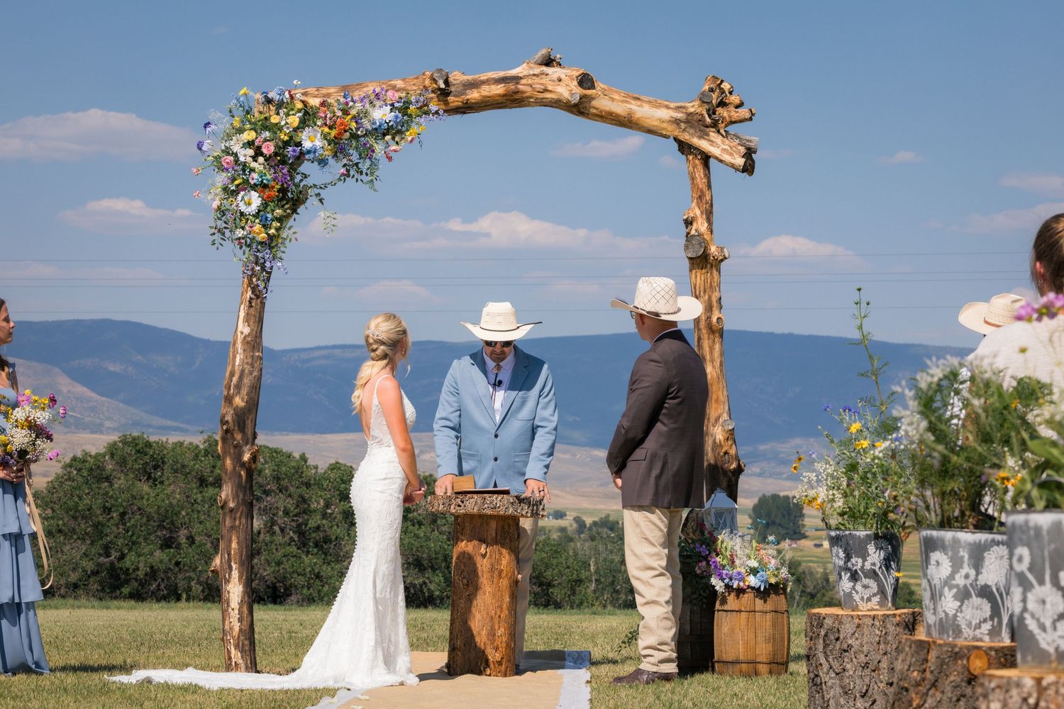 Outdoor rustic wedding ceremony under a wooden floral arch with mountain views and rolling green hills in the background.