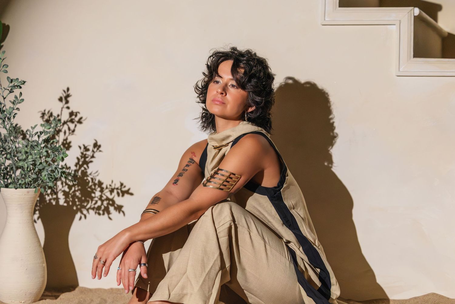 Woman with curly hair and arm tattoos sits on steps in beige outfit, posed against a neutral wall with plant.