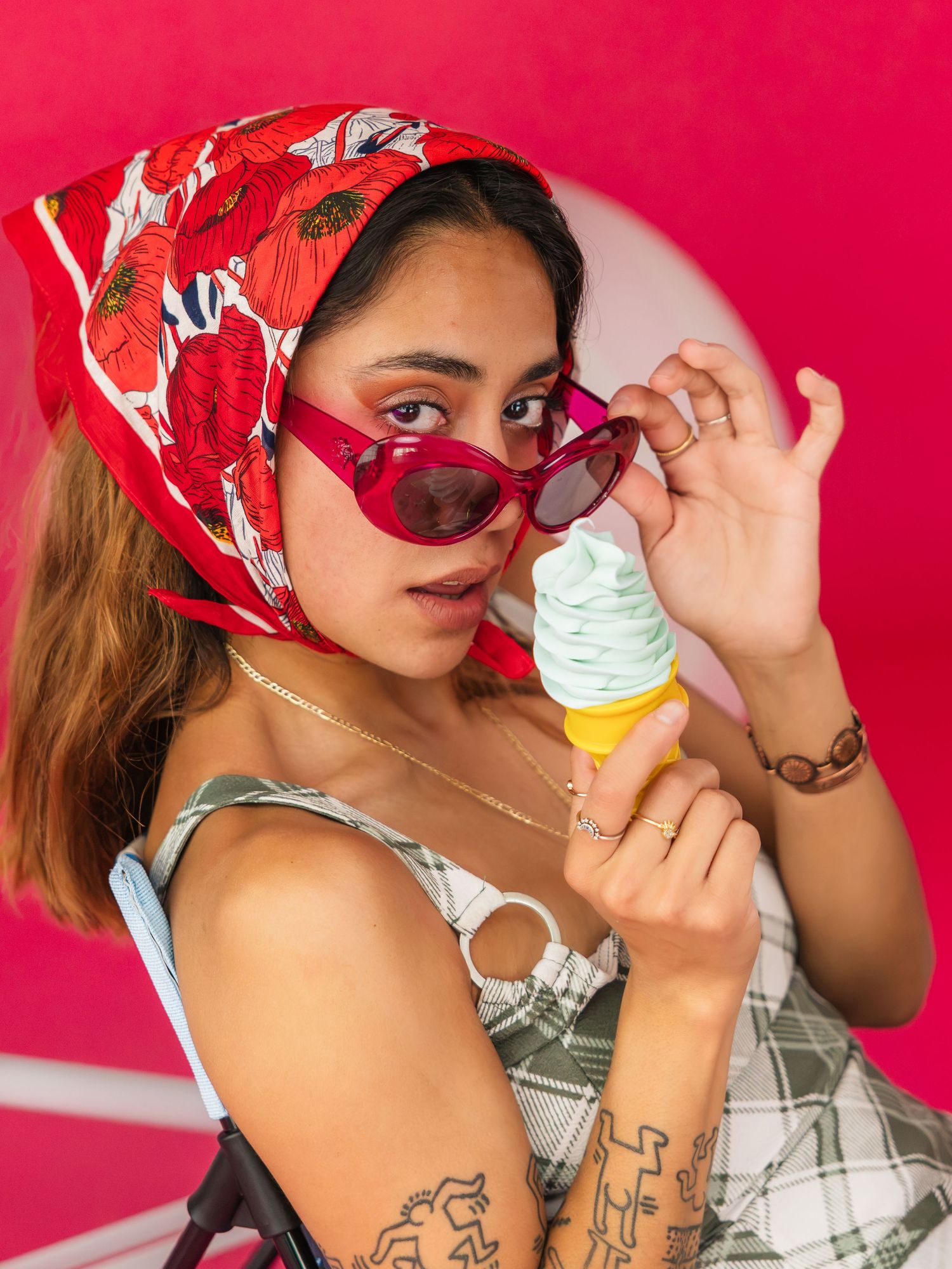 Young woman with red sunglasses, floral headscarf, and tattoos holding a mint soft serve ice cream cone.