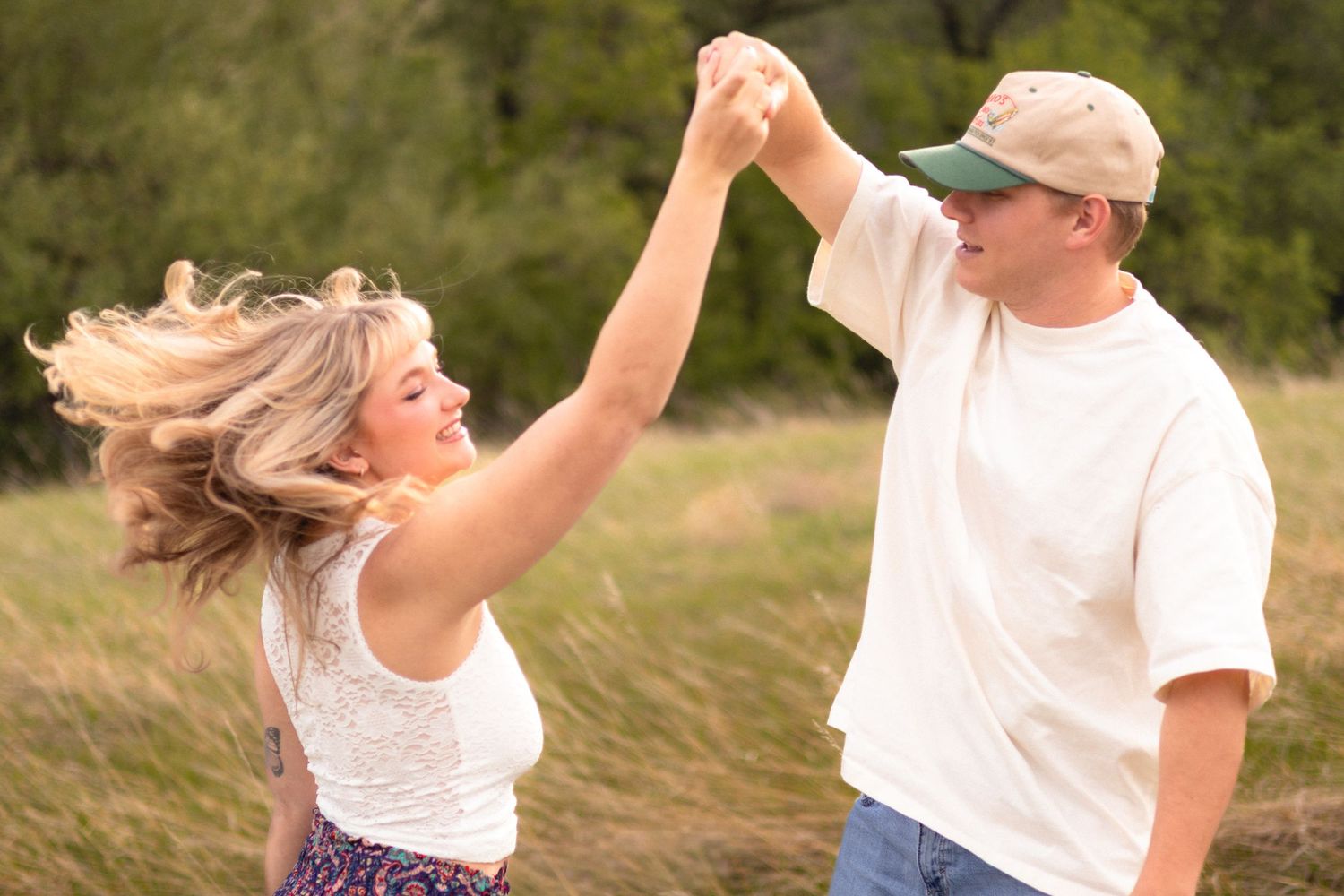 A couple dances joyfully in a grassy field, the woman spinning with flowing hair as the man smiles in a cap.