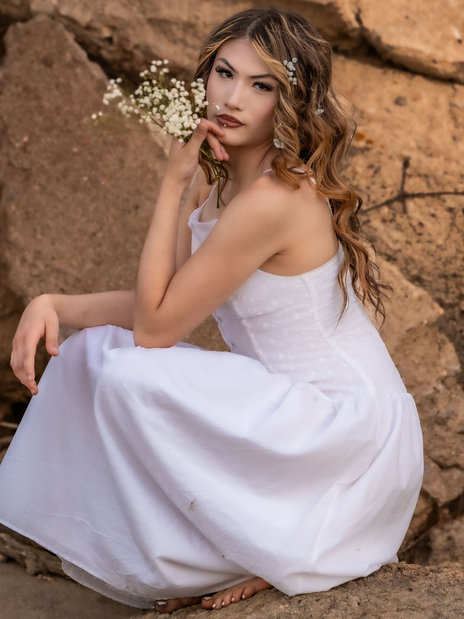 Elegant young woman in white gown holding baby's breath flowers seated on rocks in Littleton, Colorado.