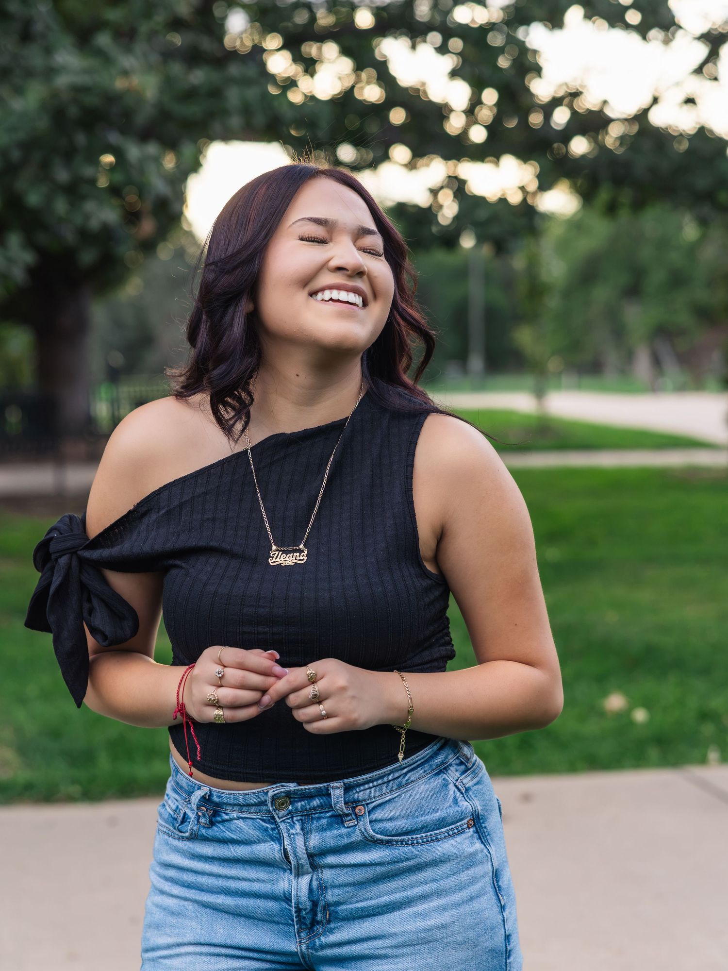 Young woman laughing joyfully outdoors, wearing black off-shoulder top and jeans, gold necklace, park background.