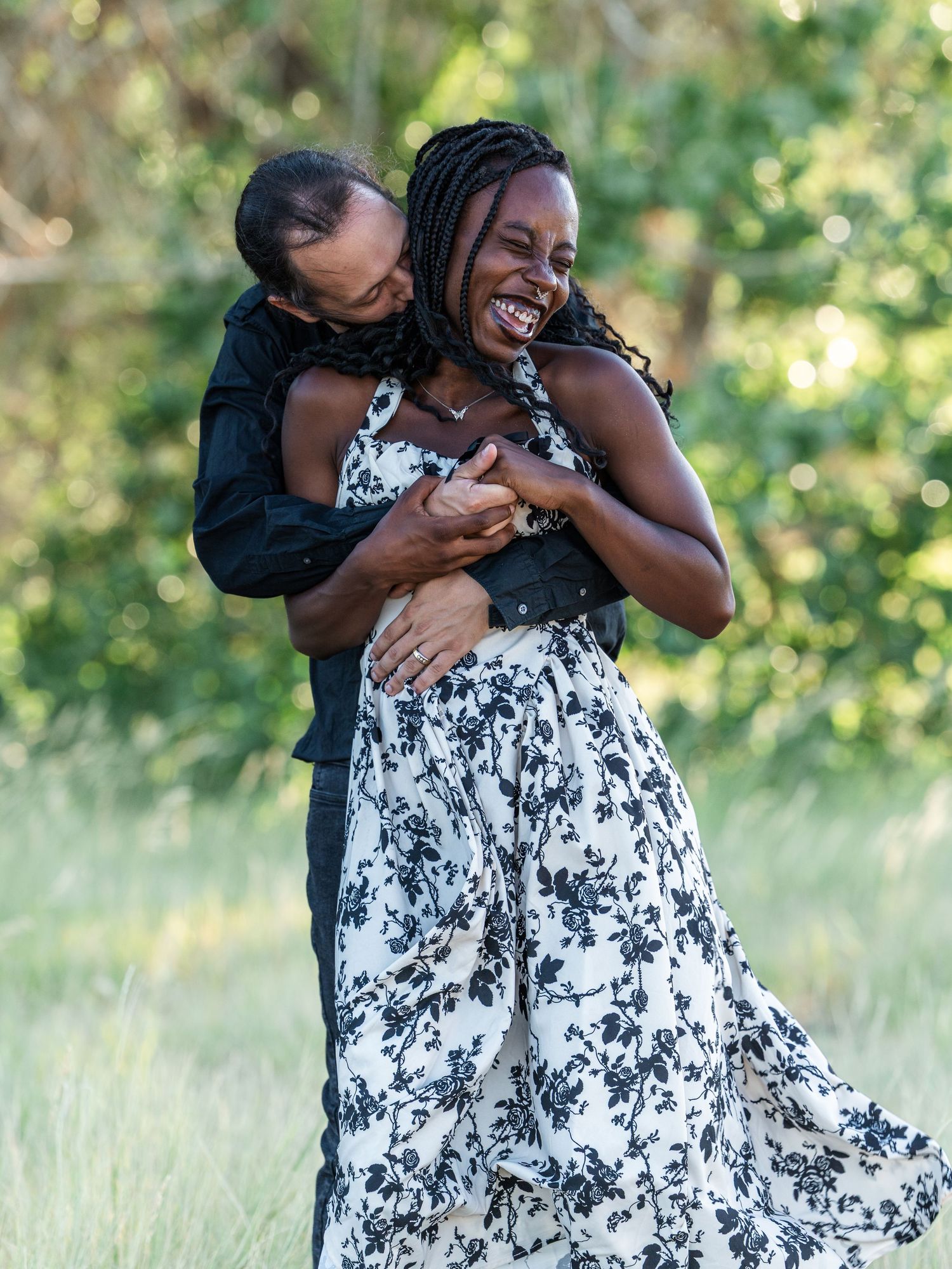 A joyful Black couple embraces and laughs together outdoors in a lush green field, woman wearing floral dress.