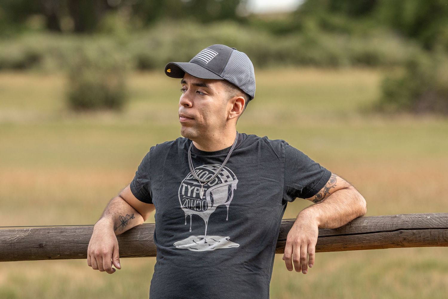 Man in gray cap and dark graphic tee leaning on wooden fence rail in outdoor field setting.