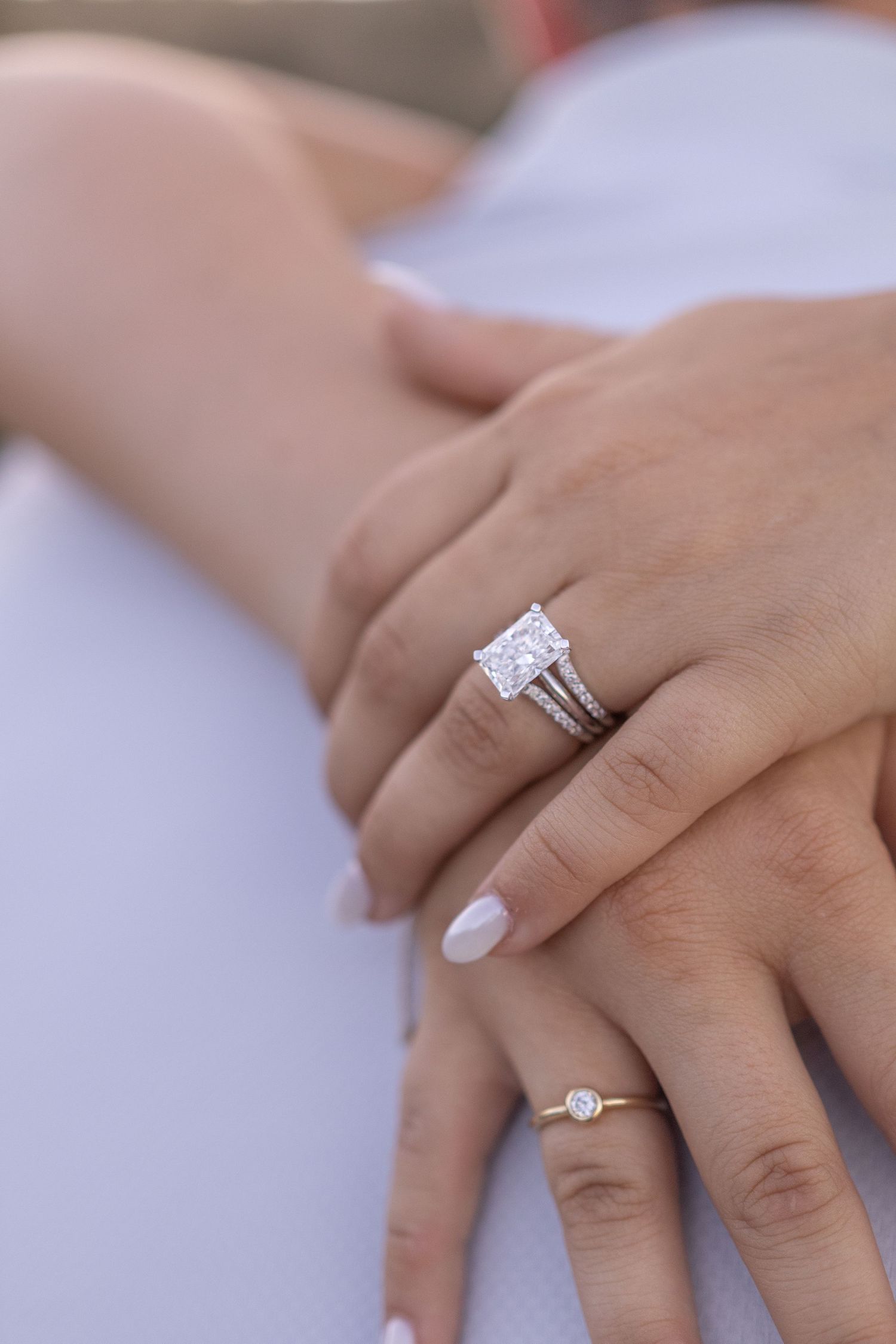 Close-up of two hands wearing diamond engagement rings, one featuring a large radiant-cut solitaire in a split-shank setting.