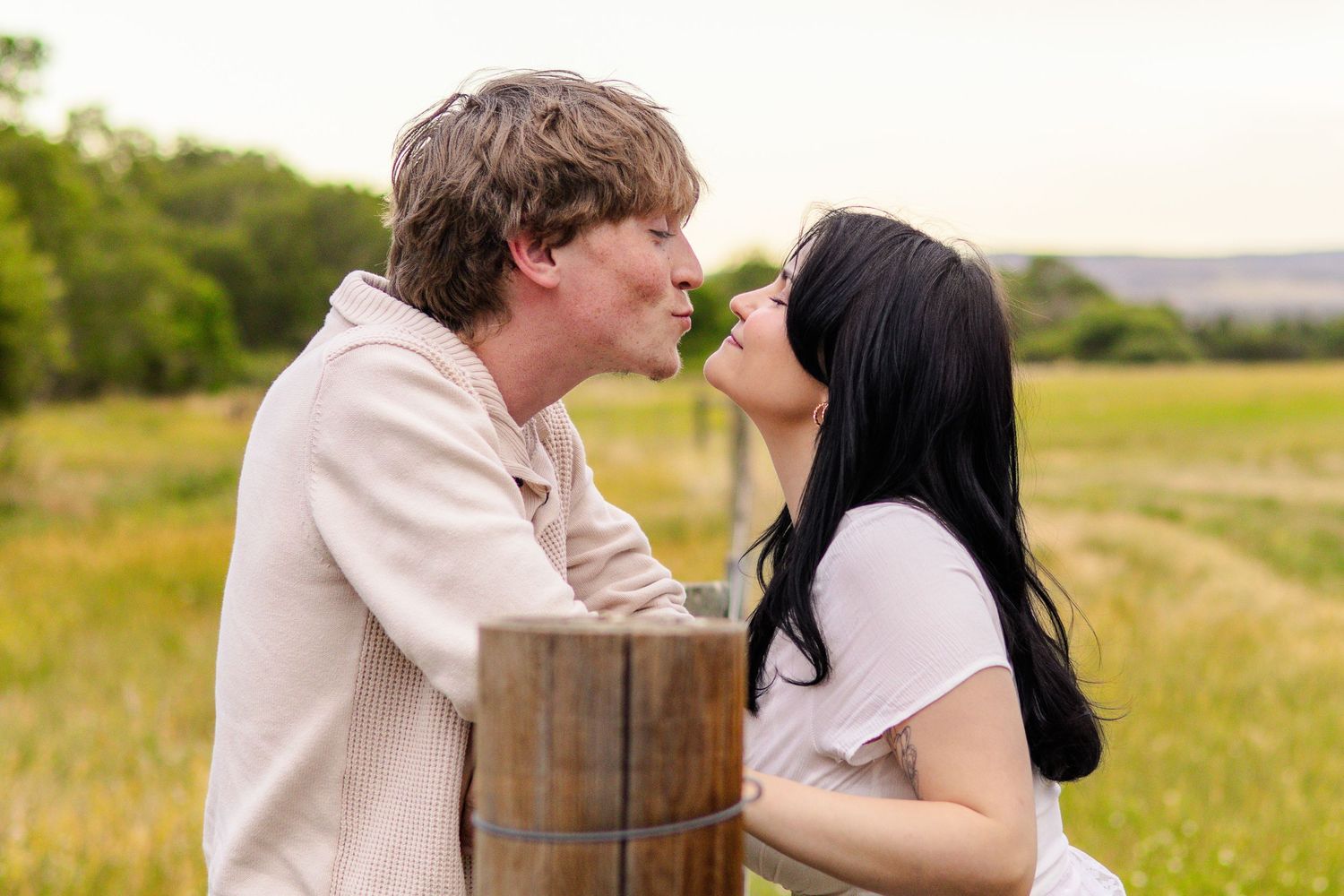 A couple shares a romantic moment about to kiss near a wooden post in a lush green field.