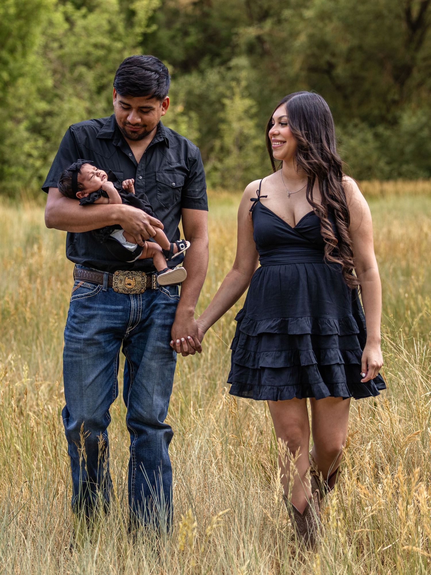 A couple in matching black outfits walks through a golden field, with the man cradling a newborn baby.