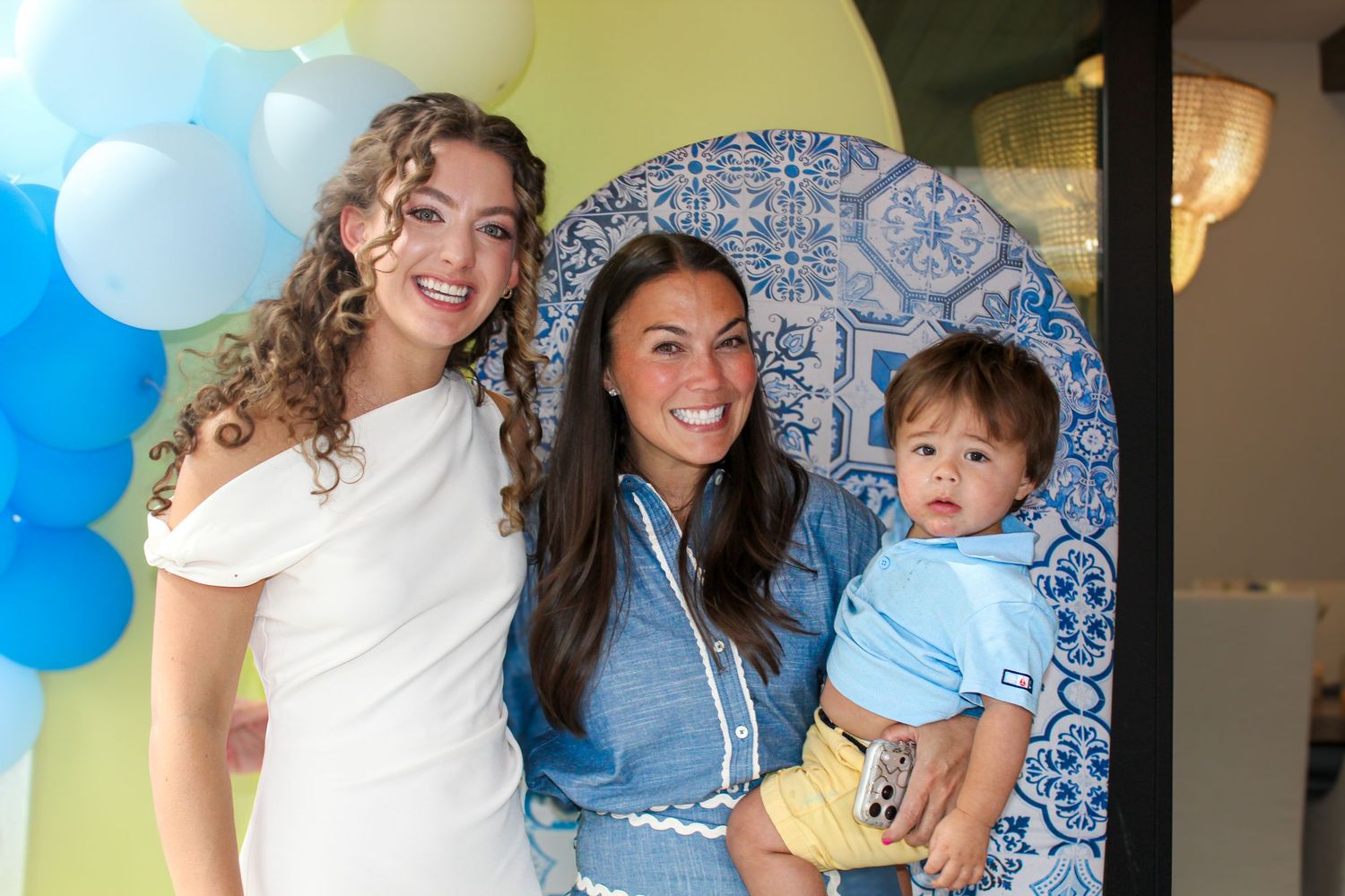 Two women and a baby boy pose together at a blue-themed party with balloon decorations.