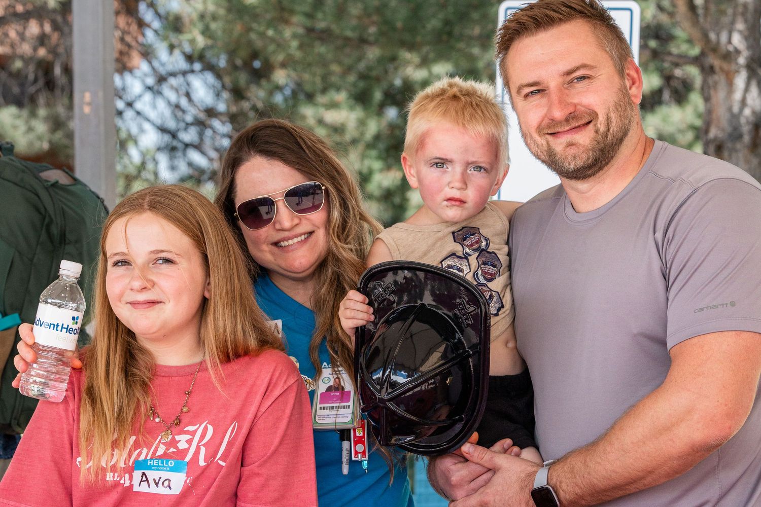Family of four smiling outdoors at community event, holding water bottle and helmet, with trees in background.