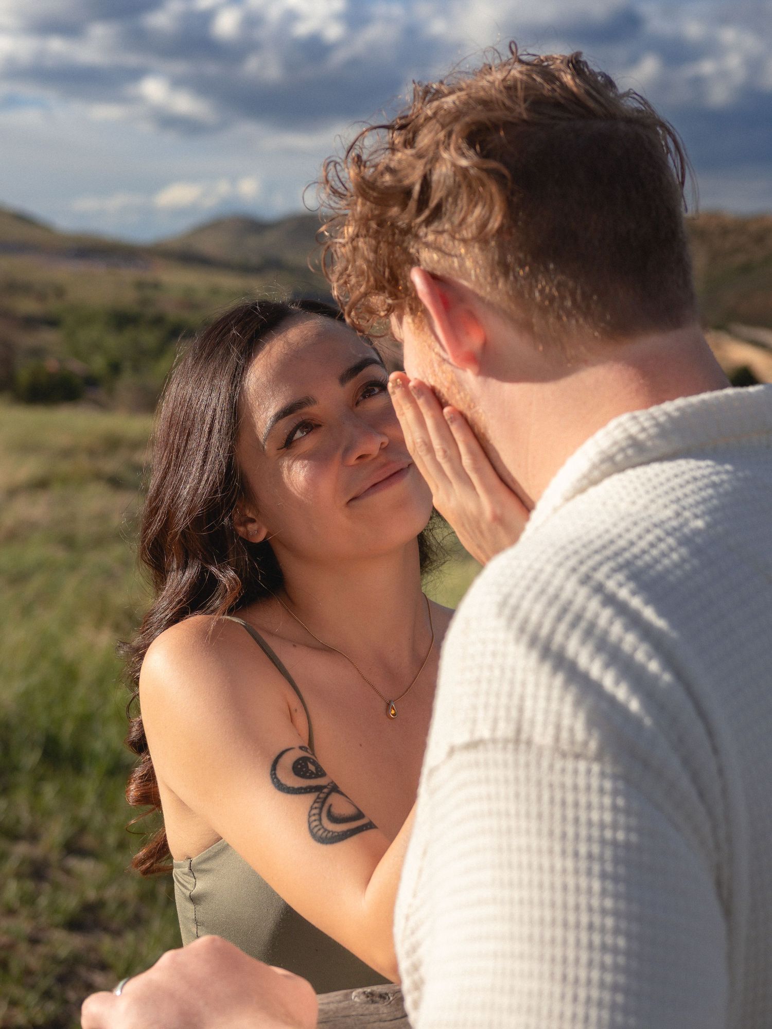 A couple shares a tender moment outdoors, with the woman smiling as the man gently cups her face.