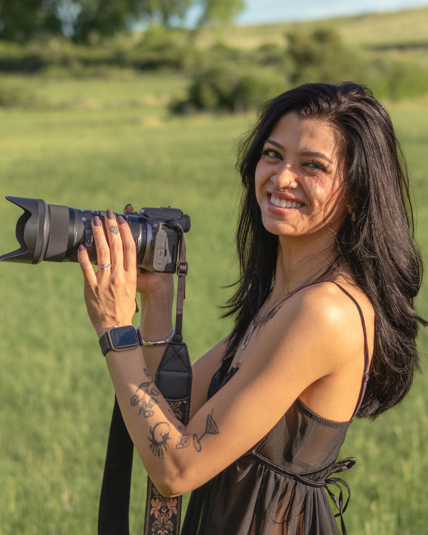 Smiling woman with long dark hair holds a DSLR camera outdoors in a green field, wearing a brown dress and tattoos.