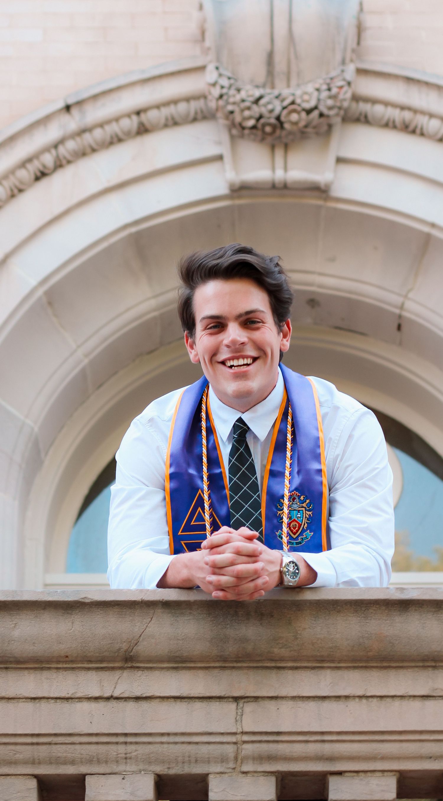 Smiling young man in graduation stole leans on stone balcony railing of classic university building.