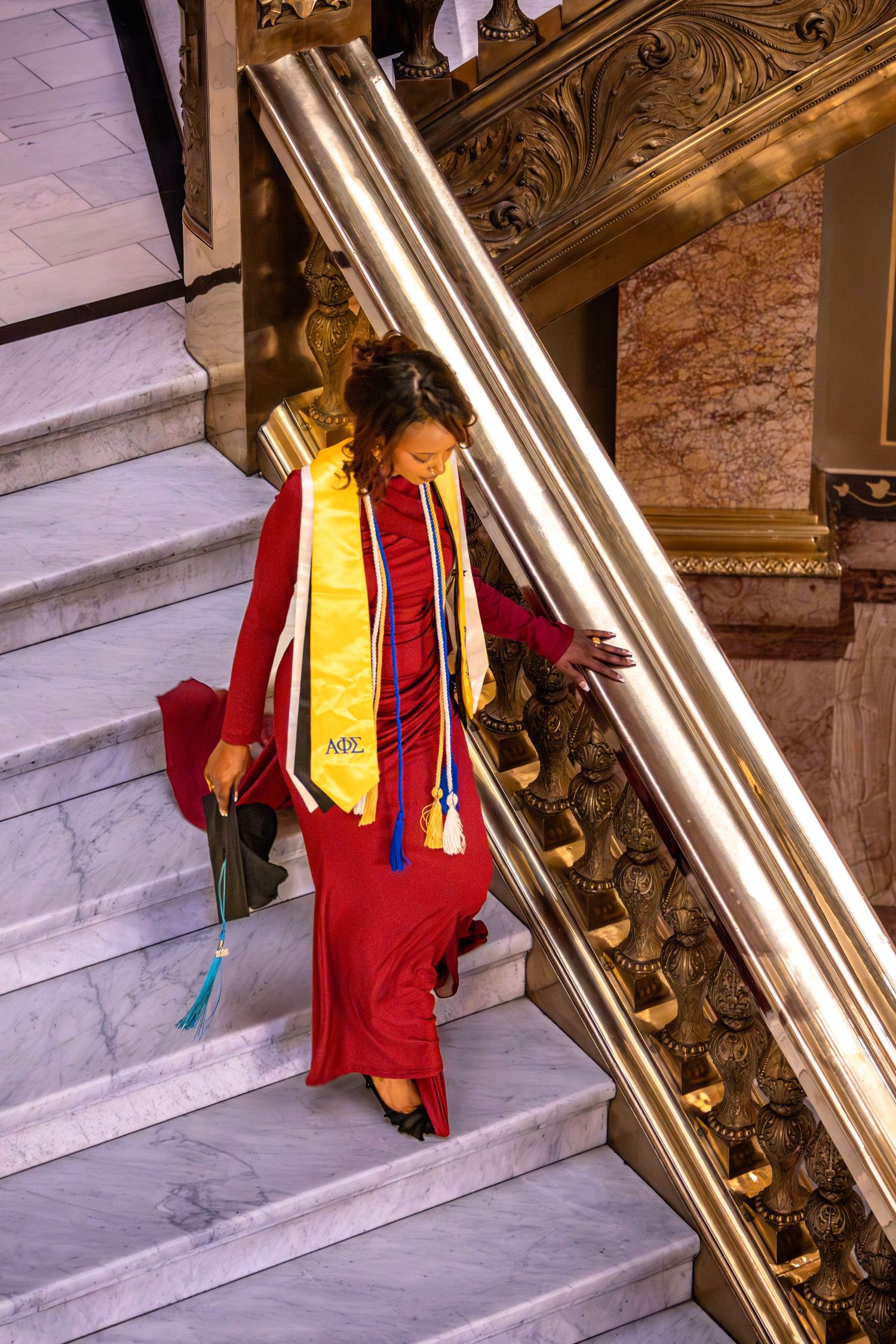 Graduate in red dress and yellow stole descends ornate marble staircase with gold railings holding cap.