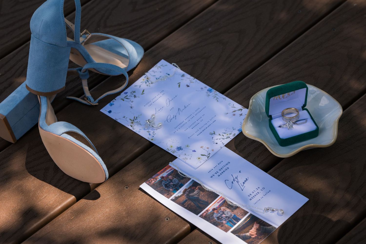 Blue wedding heels, ceremony program, photo strip, and gold rings in green velvet box arranged on wooden deck.