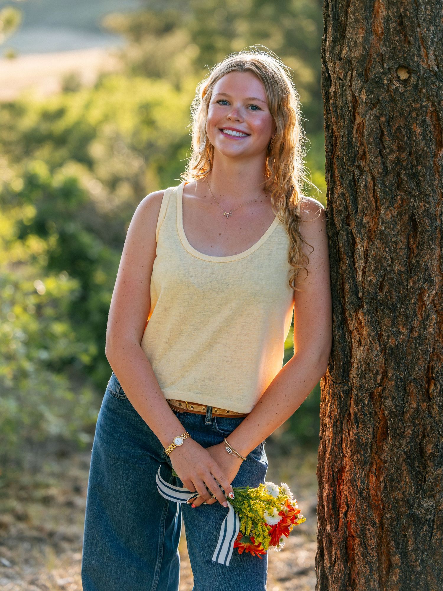 Young woman with blonde hair smiles holding wildflowers, leaning against a tree outdoors in golden light.