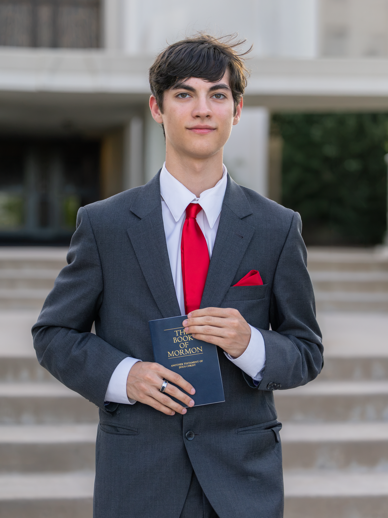 Young man in gray suit with red tie holding Book of Mormon outdoors.
