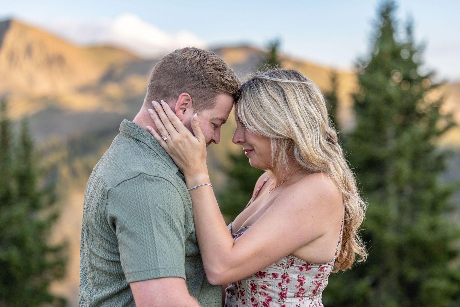 Couple sharing a romantic moment with foreheads touching, surrounded by pine trees and mountain scenery.