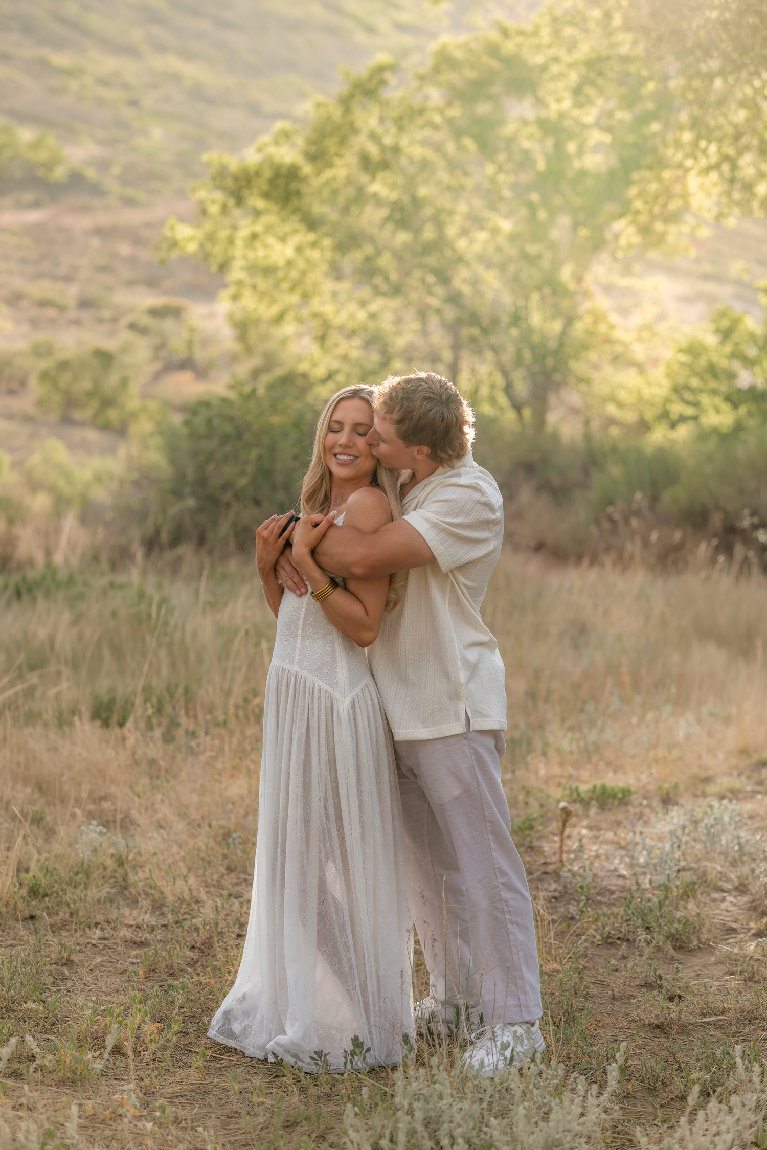 A couple embraces in a golden field at sunset, woman in white dress smiling as man kisses her cheek.
